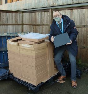 Phil standing outside in a car park, holding a laptop next to a pile of laptops in boxes on a delivery pallet. Phil is wearing a facemask. 