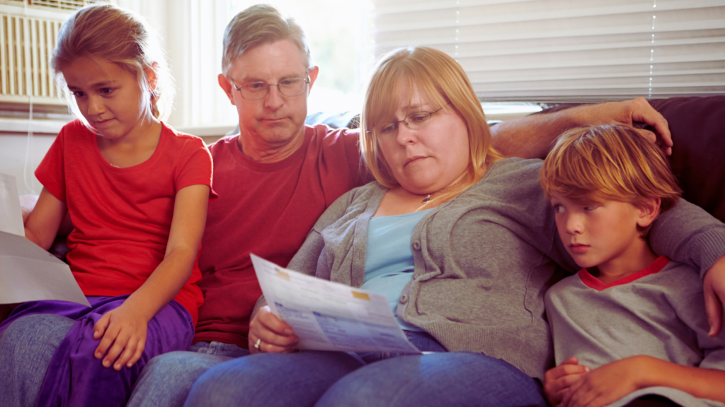 A family with two adults and two children sat on a sofa looking unhappy, the woman has a bill in her hand.