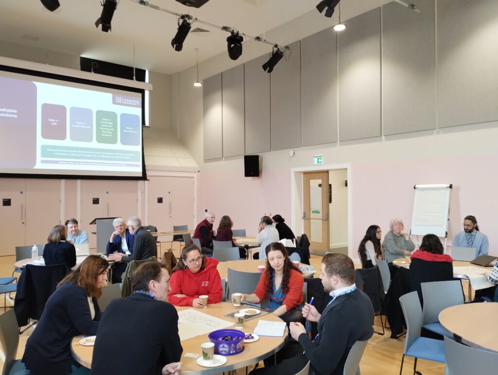 People sat in a hall around round tables in deep discussion. We see a Powerpoint projected behind them on a big screen.