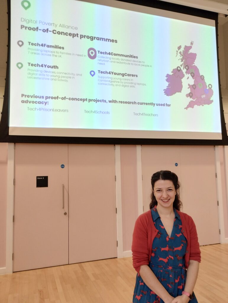 Immy from Digital Poverty Alliance standing in front of a big screen that is projecting a Powerpoint presentation. She is wearing a blue dress and has long brown hair.