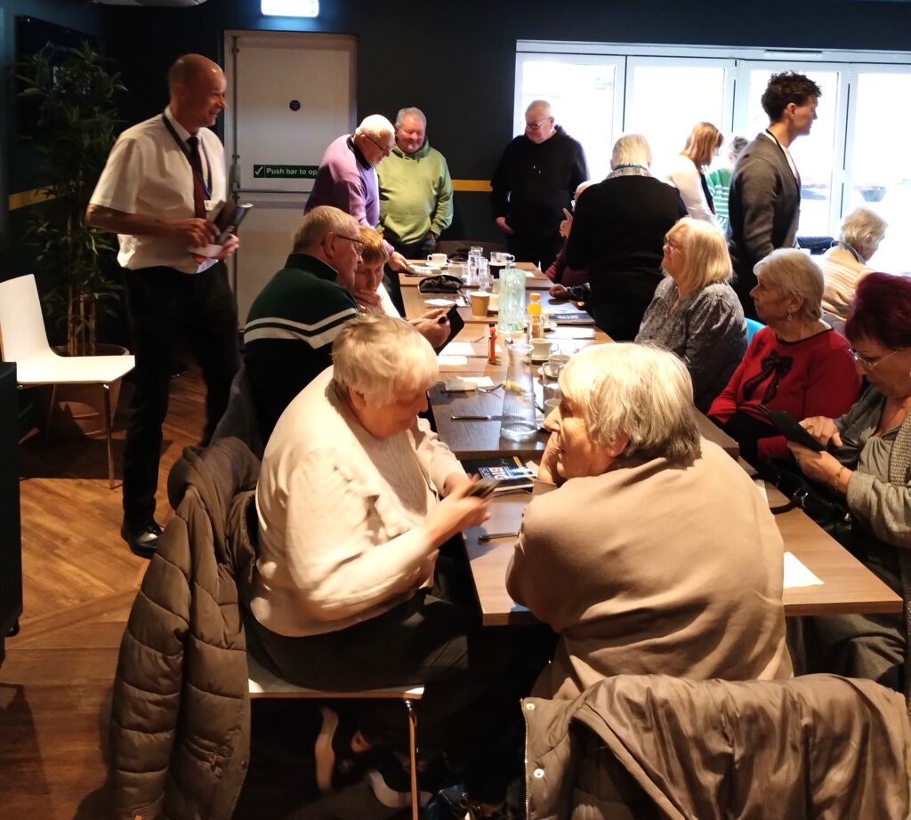 People sat round tables in a hall chatting with others handing out leaflets