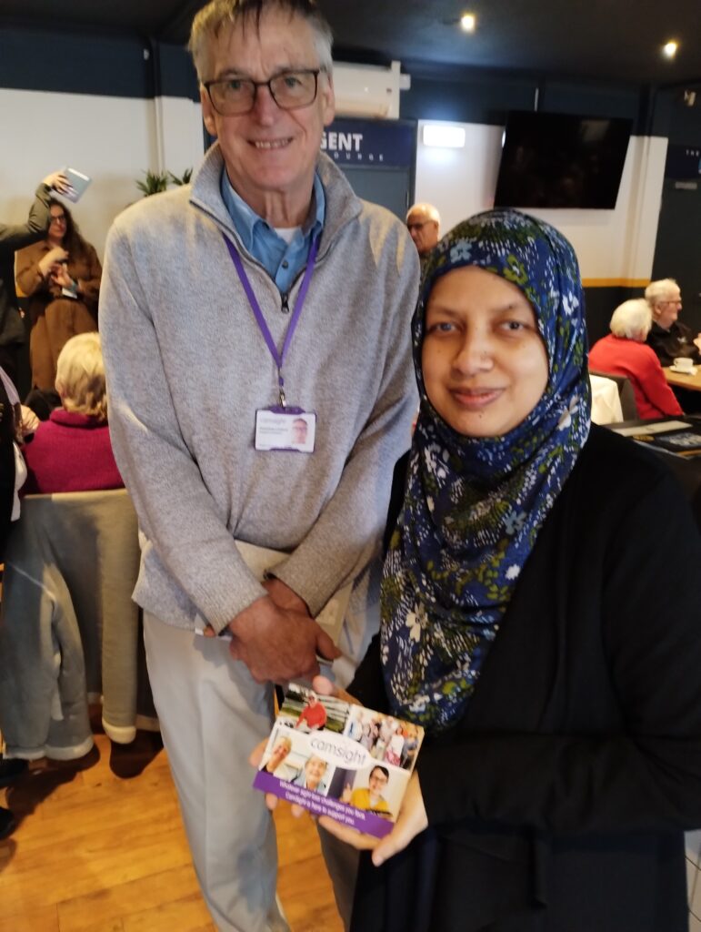 Two people smile to camera. A man is wearing glasses and a grey jumper. The woman is wearing a black dress and hijab. She holds a CamSight leaflet.