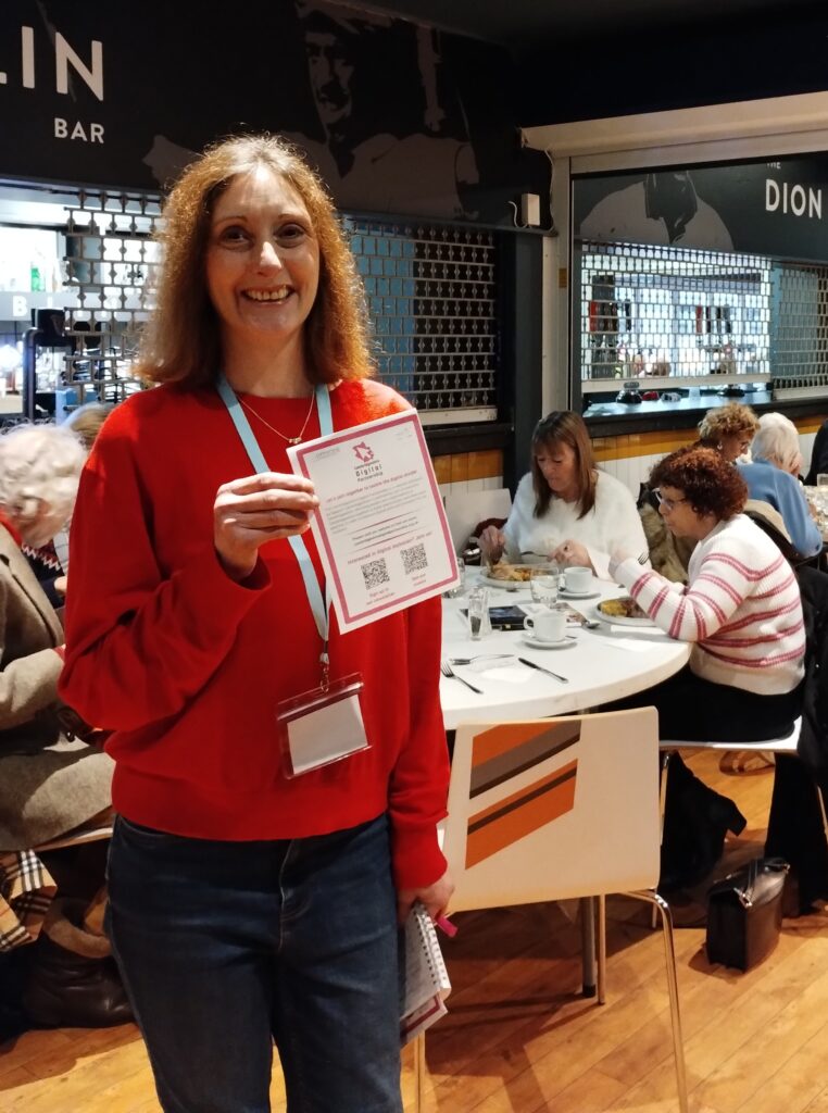 A woman in a red jumper holds up a CDP leaflet. People are eating lunch behind her.